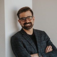 Man in glasses smiling while leaning against a wall